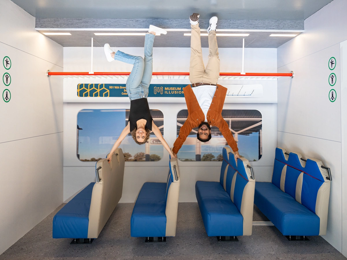 A smiling man and woman defying gravity in the Reversed Room in Museum of Illusions Denver. The room represents the interior of the Denver passenger train.